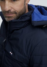 Jachetă puffer de culoare bleumarin cu căptușeală interioară albastră, având o textură matlasată, guler înalt, închidere cu fermoar și accente cu logo pe piept.