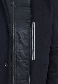 Chaqueta de marina que combina lana y tejido acolchado, con cremallera frontal, botones y un detalle de bolsillo en contraste. Textura suave y corte entallado.