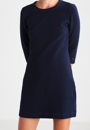 Woman wearing a navy blue, long-sleeve, knee-length, casual dress with a round neckline and a textured fabric.