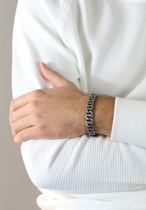 Metal bracelet with a woven design in silver and black tones, worn on a wrist covered by a textured white long-sleeve shirt.
