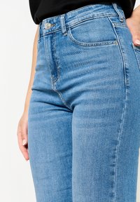 Jeans de mezclilla azul de tiro alto con una textura suave, que cuentan con un bolsillo frontal, cierre de botón metálico y detalles de costura visibles.