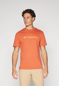 Narančasta pamučna majica s okruglim izrezom, sadrži žuti Columbia logo na prednjoj strani. Model nosi svijetlosmeđe šortseve.
