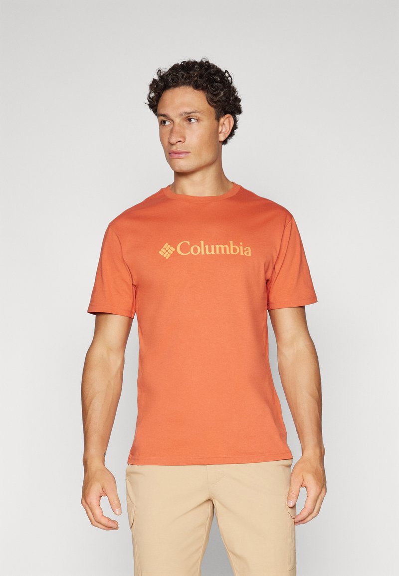 Narančasta pamučna majica s okruglim izrezom, sadrži žuti Columbia logo na prednjoj strani. Model nosi svijetlosmeđe šortseve.