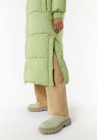 Padded długa kurtka w jasnym zielonym kolorze z detalami w postaci zatrzasków, w połączeniu z szerokimi beżowymi spodniami i masywnymi jasnobrązowymi botkami.
