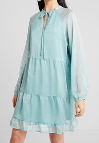 Robe bleu clair à motif à pois, design à volants, manches longues transparentes et encolure fendue avec un lien à nouer.