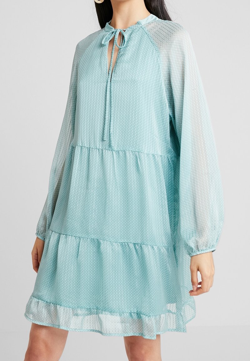 Robe bleu clair à motif à pois, design à volants, manches longues transparentes et encolure fendue avec un lien à nouer.