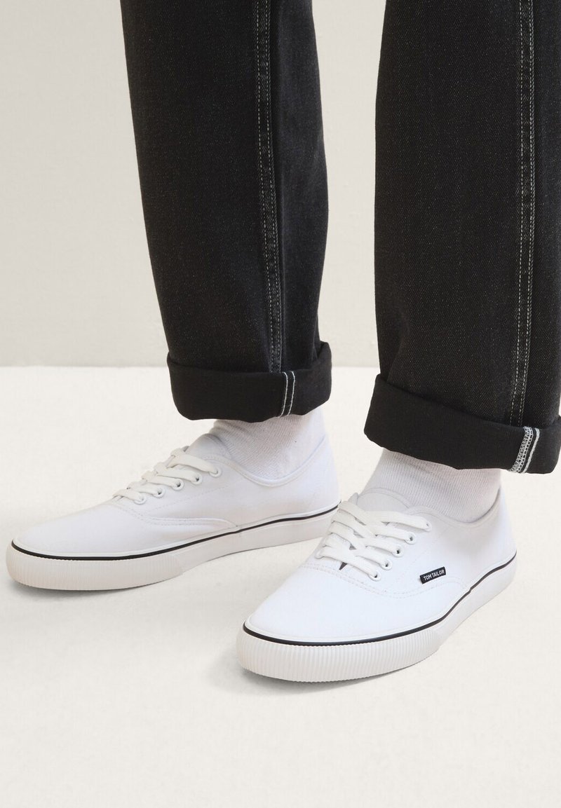 Sneaker di tela bianchi con dettagli neri, punta rotonda, suola piatta in gomma e sei fori per i lacci, indossati con pantaloni neri arrotolati e calze bianche.