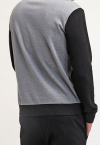 Sweat-shirt gris et noir avec des manches texturées, des poignets côtelés et une coupe décontractée. Le dos présente un design sans coutures avec des couleurs contrastantes.