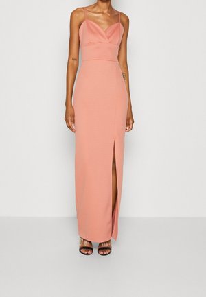 Femme portant une robe longue rose clair sans manches avec une fente sur la cuisse et des bretelles spaghetti, associée à des talons ouverts noirs.