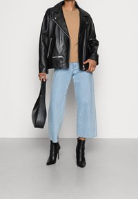 Schwarze Leder-Bikerjacke mit silbernen Reißverschlüssen, beiger Pullover, hellblaue Jeans mit weitem Bein und schwarze Stiefeletten. Trägt eine schwarze Tasche.