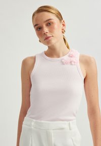 Ljust rosa, ribbad linne med rund halsringning och en blommig detalj på vänster axel. Tillverkat av mjukt, elastiskt tyg.