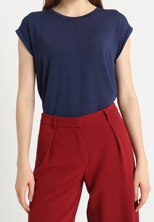 Woman wearing a navy blue short-sleeve top tucked into high-waisted, wide-leg red trousers against a plain background.