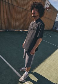 Sort kortærmet polo med lynlås og brystlomme, parret med grå shorts. Båret med grå sneakers og hvide sokker på en sportsbane.
