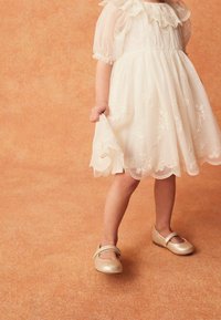 Robe blanche brodée avec manches bouffantes, ourlet festonné et surcouche transparente. Chaussures Mary Jane dorées avec une bride, sur un fond orange.
