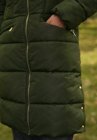Zelená prešívaná bunda s kapucňou, s predným zipsom, bočnými vreckami a zlatými detailmi na gombíkoch na spodnom okraji. Textúra sa zdá byť hladká.