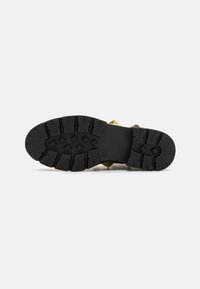 Suela de zapato de goma negra con un diseño de surcos profundos, que presenta dos tachuelas de pirámide doradas cerca del talón. Superficie suave con patrones de agarre texturizados.