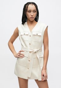 Κρεμ αμάνικο φόρεμα με ντεκολτέ σε σχήμα V, διπλές τσέπες στο στήθος, χρυσά κουμπιά και ζώνη στη μέση. Υφασμάτινη υφή με κοντό μήκος.