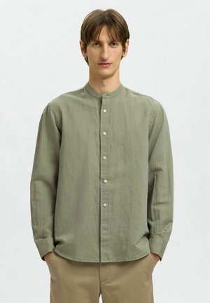 Jeune homme debout, les mains dans les poches, portant une chemise boutonnée col mao vert olive clair et un pantalon beige sur un fond uni.