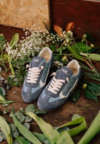 Paire de baskets P448 bleu et gris avec des lacets blancs posées sur un sol en bois entourées de feuilles vertes et de petites fleurs blanches.
