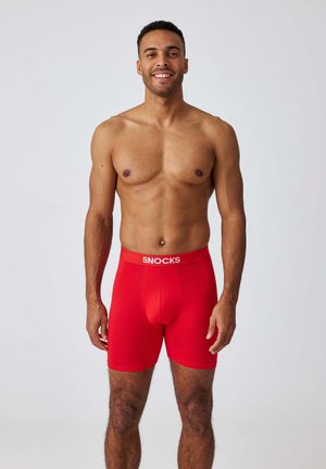 Glunderende, shirtloze man staat frontaal en draagt rode boxershorts met het "SNOCKS"-logo op de tailleband, tegen een effen witte achtergrond.