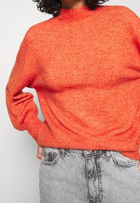 Jasnopomarańczowy sweter dzianinowy z wysokim kołnierzem, luźnym krojem i ściągaczami. Noszony z szarymi dżinsami o klasycznym pięciokieszeniowym fasonie.