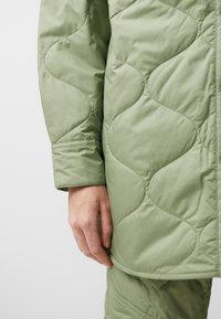 Jachetă pufosă de un verde deschis, cu un model ondulat, având închidere cu nasturi și mâneci cu mansete, prezentată cu o mână sprijinită pe material.