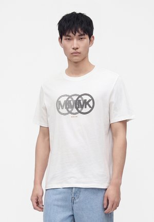 Ung mand med kort sort hår iført en hvid t-shirt med en sortstripet cirkulær logo og lyseblå jeans, stående foran en ensfarvet baggrund.