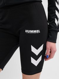 Zwarte sportshorts met wit Hummel-logo en dubbel chevron-patroon op de dij, gedragen door een persoon met blauwe nagellak op de vingernagels.