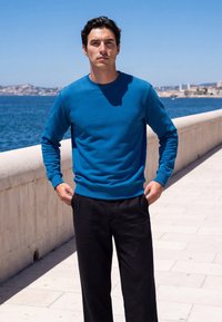 Man in blue long-sleeve shirt and black pants standing on a seaside promenade with ocean and distant cityscape in the background.