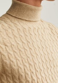 Pull beige en maille torsadée avec col roulé porté par une personne, montrant la texture détaillée et le col côtelé.