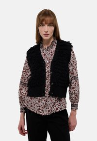Gilet en tricot noir avec des détails en volants texturés, porté par-dessus une chemise à motif floral rouge et blanc, présentant un col rond et des boutons.