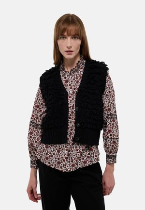 Gilet en tricot noir avec des détails en volants texturés, porté par-dessus une chemise à motif floral rouge et blanc, présentant un col rond et des boutons.