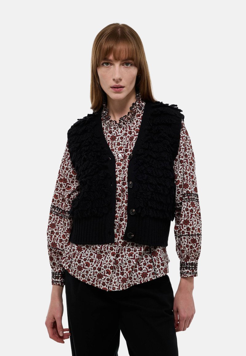 Gilet en tricot noir avec des détails en volants texturés, porté par-dessus une chemise à motif floral rouge et blanc, présentant un col rond et des boutons.