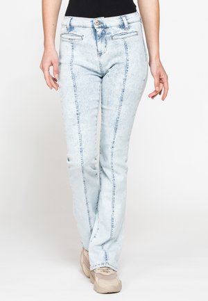 Donna che indossa jeans azzurri chiaro lavaggio acido con cuciture verticali lungo le gambe e sneakers beige, in piedi davanti a uno sfondo bianco uniforme.