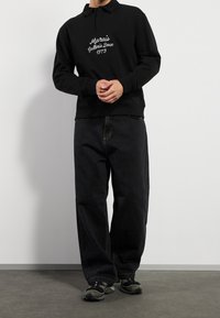 Sort sweatshirt med polo-krave, med hvidt broderet tekst. Båret med løse sorte jeans og sorte atletiksko.