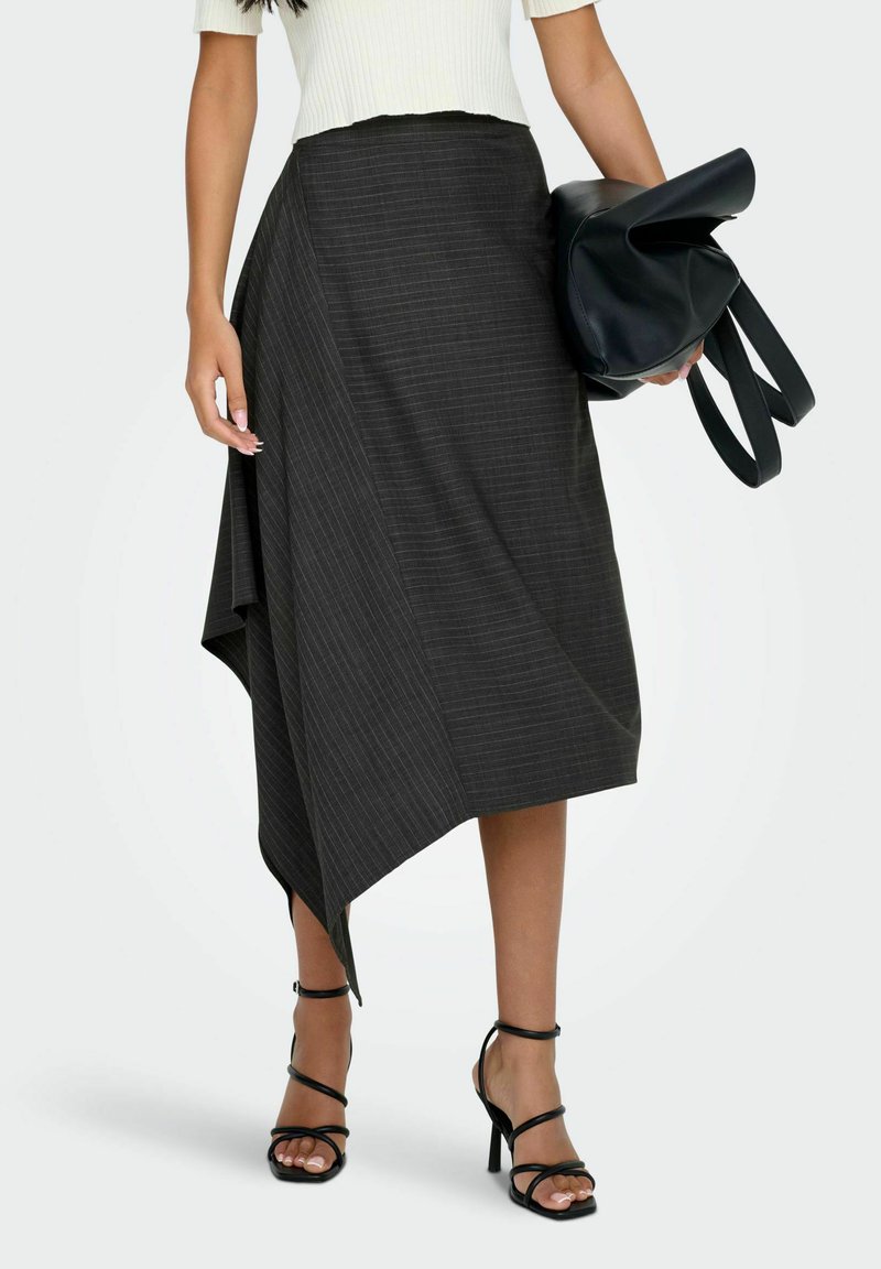 Vrouw in een asymmetrische zwarte gestreepte rok, witte korte mouwen top, zwarte strappy hakken, met een gevouwen zwarte leren tas.