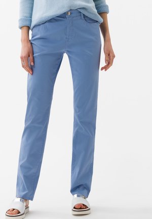 Osoba nosząca jasnoniebieskie spodnie o prostych nogawkach, blado-niebieski sweter o fakturze, srebrne sandały na płaskiej podeszwie z odkrytymi palcami, stojąca na białym tle.