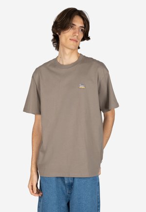Young man with medium-length brown hair wearing a loose brown t-shirt with a small camper van graphic and blue denim shorts.