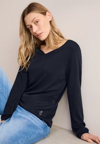 Maglietta a maniche lunghe blu navy con scollatura a V, realizzata in tessuto morbido, con un logo in evidenza sull'orlo, abbinata a jeans azzurri.