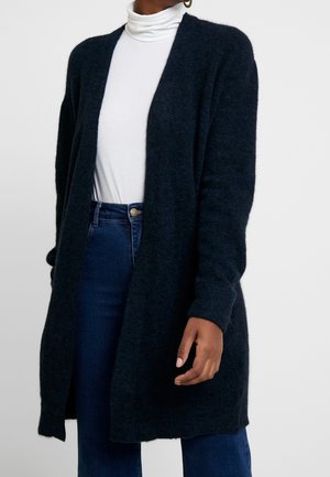 Femme portant un long cardigan noir sur un col roulé blanc et un jean bleu taille haute, debout devant un fond uni.