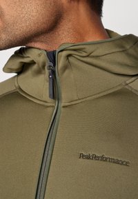 Olivengrøn hættejakke med lynlås, lavet af blødt, tekstureret stof, med broderet "PeakPerformance" logo på brystet.