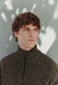 Gebreide trui met hoge kraag in donkerbruin met een gestructureerd patroon en zwarte rits. Het model heeft warrig bruin haar en een lichte huid.