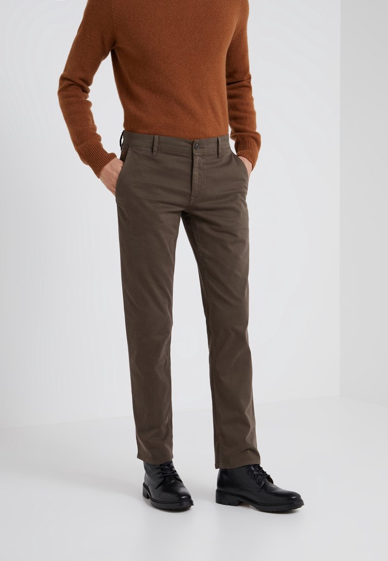 Homme portant un pull marron, un pantalon marron foncé et des bottes noires à lacets, debout les mains dans les poches devant un fond uni.