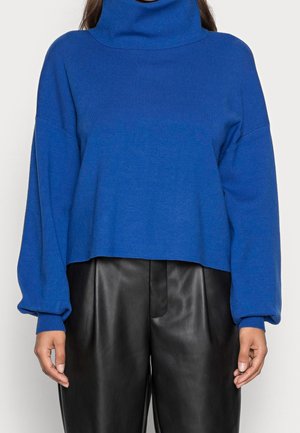 Femme portant un pull à col roulé à manches longues bleu et un pantalon en cuir taille haute noir, debout les bras détendus.