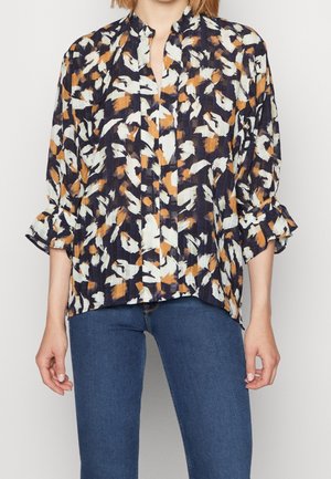 Kvinde iført en løs, langærmet bluse med flæsekant og abstrakt sort, hvid og orange mønster, kombineret med blå jeans.