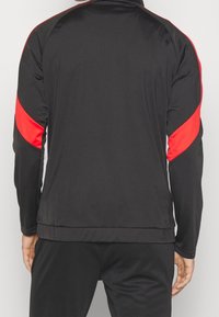 Schwarze Trainingsjacke mit langen Ärmeln, die rote Akzente an den Schultern und ein glattes, feuchtigkeitsableitendes Material aufweist.