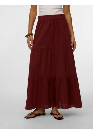 Femme portant une longue jupe bordeaux à volants, un haut blanc, des bracelets en bois et des sandales plates noires, debout sur un sol gris clair.