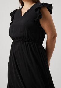 Kvinna iklädd en svart klänning med fladdrande ärmar och midja som är åtdragen, poserande mot en enkel ljus bakgrund.