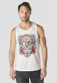 Hombre con camiseta sin mangas blanca con estampado colorido de calaveras tropicales, pájaros y flores, y pantalones negros, de pie contra un fondo gris liso.