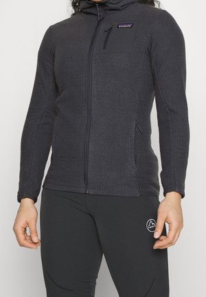 Personne portant une veste zippée noire texturée Patagonia avec une poche poitrine et un pantalon noir La Sportiva avec logo sur la cuisse.